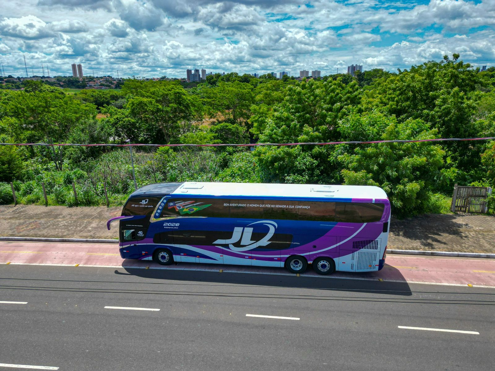 Ônibus da JL Expresso percorrendo rodovia com a cidade ao fundo, em vista aérea